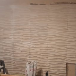 Arkadiusz Pawlak - Montaż paneli ściennych 3D w trakcie prac wykończeniowych, widoczne niedociągnięcia przy krawędziach i otwór na instalację, drabina i worek z klejem w tle.