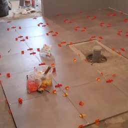 Arkadiusz Pawlak - Układanie szarych płytek podłogowych z użyciem pomarańczowych klipsów poziomujących; widoczny grzejnik elektryczny, narzędzia i nieukończone fragmenty podłogi.