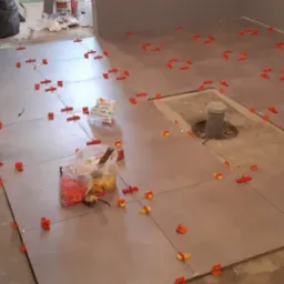 Układanie szarych płytek podłogowych z użyciem pomarańczowych klipsów poziomujących; widoczny grzejnik elektryczny, narzędzia i nieukończone fragmenty podłogi.