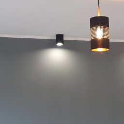 Arkadiusz Pawlak - Szara ściana oświetlona trzema lampami sufitowymi punktowymi i trzema wiszącymi cylindrycznymi lampami z perforacją i złotym wnętrzem.