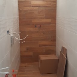 Arkadiusz Pawlak - Wnętrze łazienki w trakcie wykańczania z ułożonymi płytkami imitującymi drewno na podłodze i ścianie akcentującej, białe płytki na pozostałych ścianach, widoczne wystające kable i rury oraz kartony...