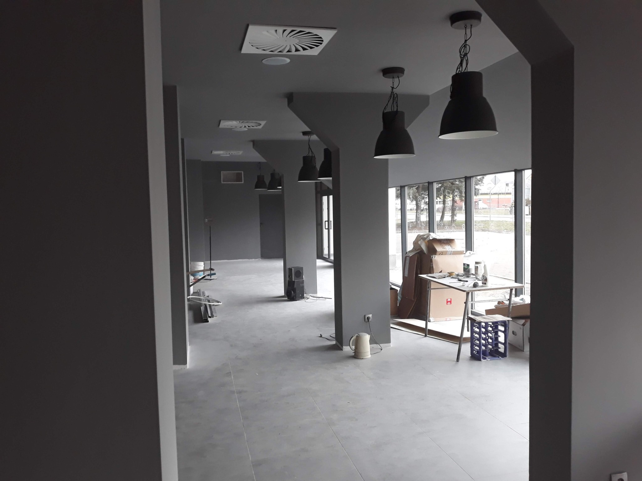 Wnętrze biura w trakcie remontu: szare ściany, betonowa podłoga, czarne lampy wiszące, kartony, stół z narzędziami, czajnik elektryczny i głośnik na podłodze.