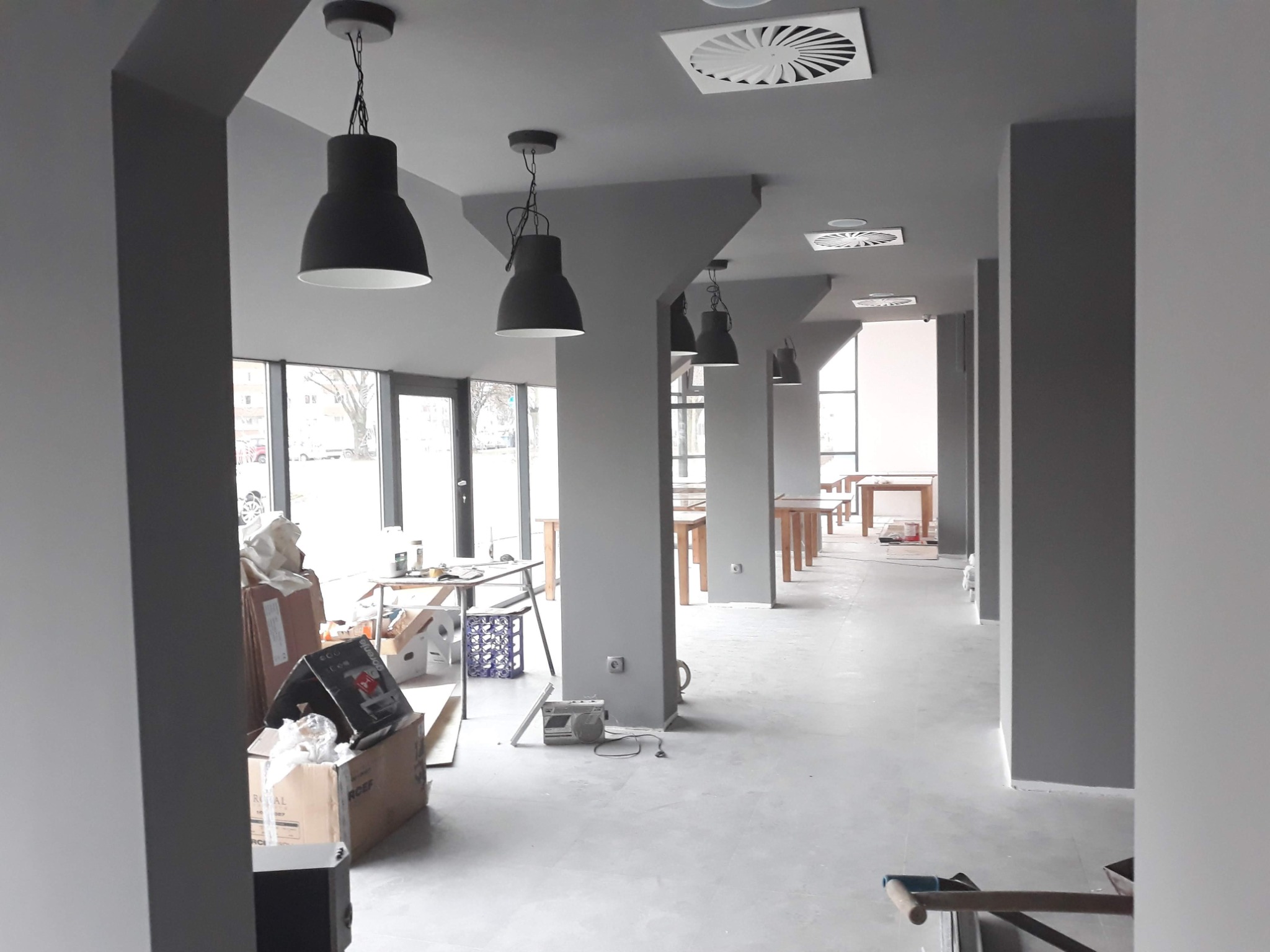 Wnętrze biura w Opalenicy w trakcie remontu, świeżo pomalowane na szaro, widoczne kartony, lampy wiszące i prowizoryczne stoły.