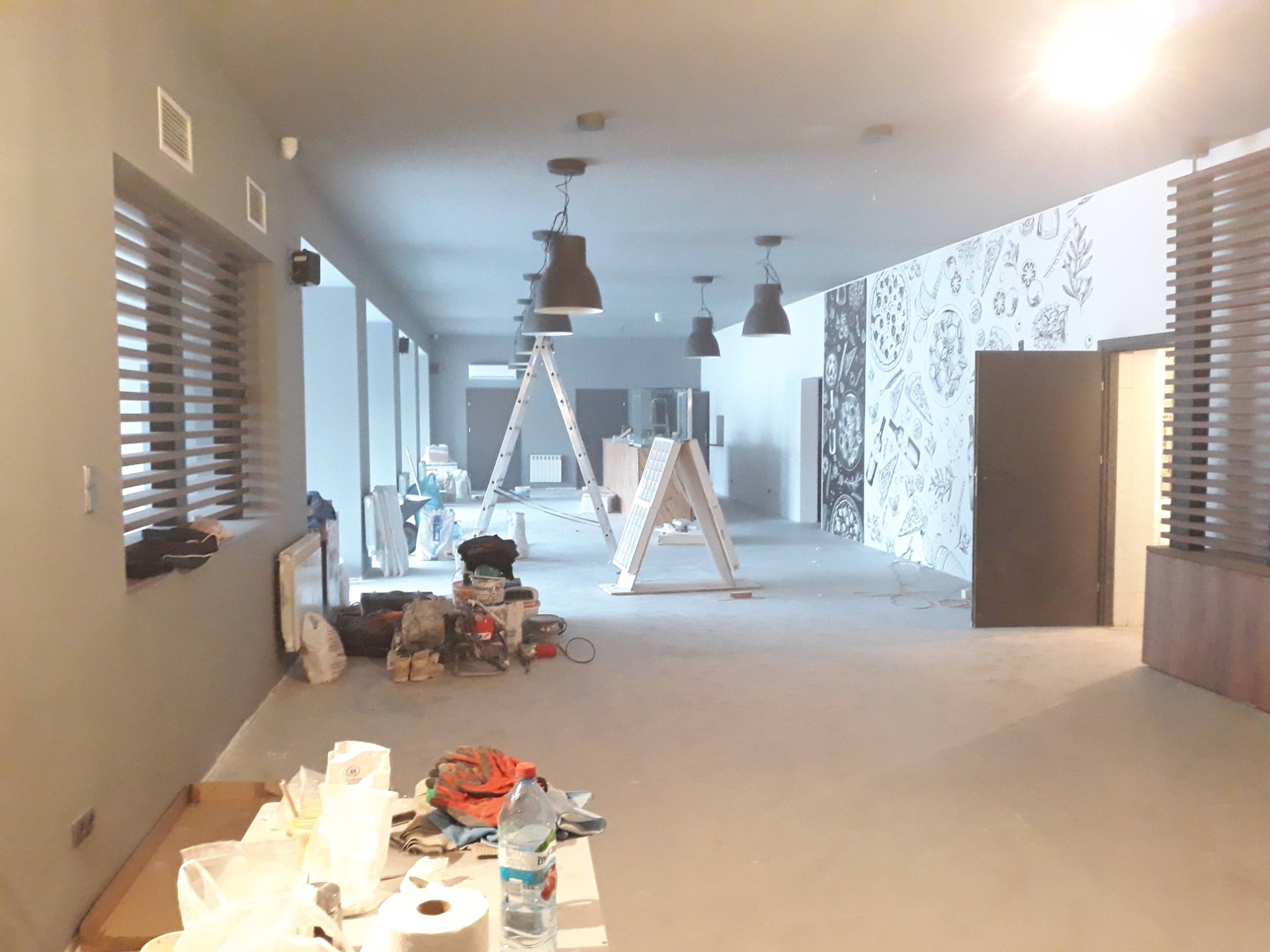 Przestronne wnętrze w trakcie remontu: widoczne drabiny, narzędzia, materiały budowlane i dekoracyjna grafika na ścianie, sugerujące etap przygotowawczy do wykończenia.