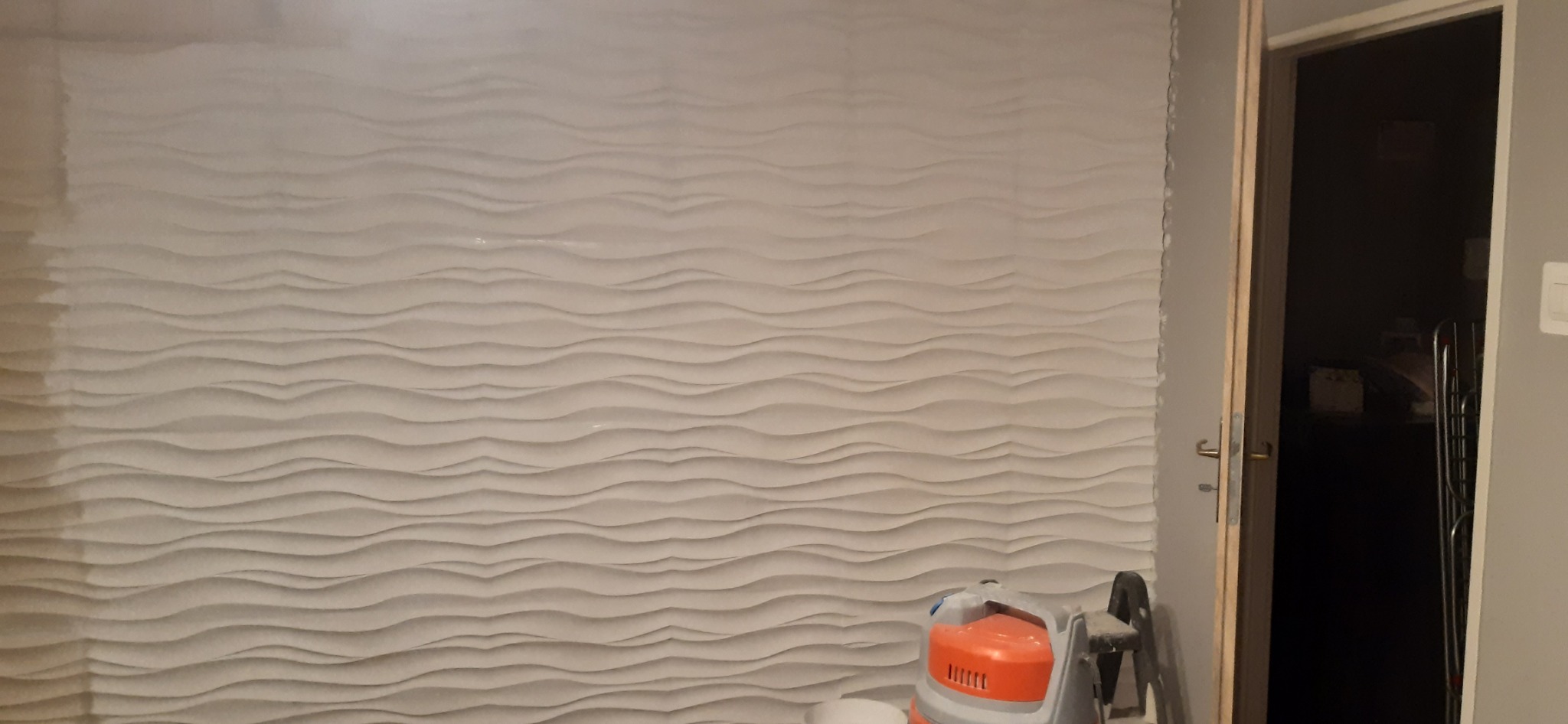 Ściana pokryta dekoracyjnymi panelami 3D w odcieniach bieli, z widocznym odkurzaczem przemysłowym i otwartymi drzwiami do ciemnego pomieszczenia w tle.