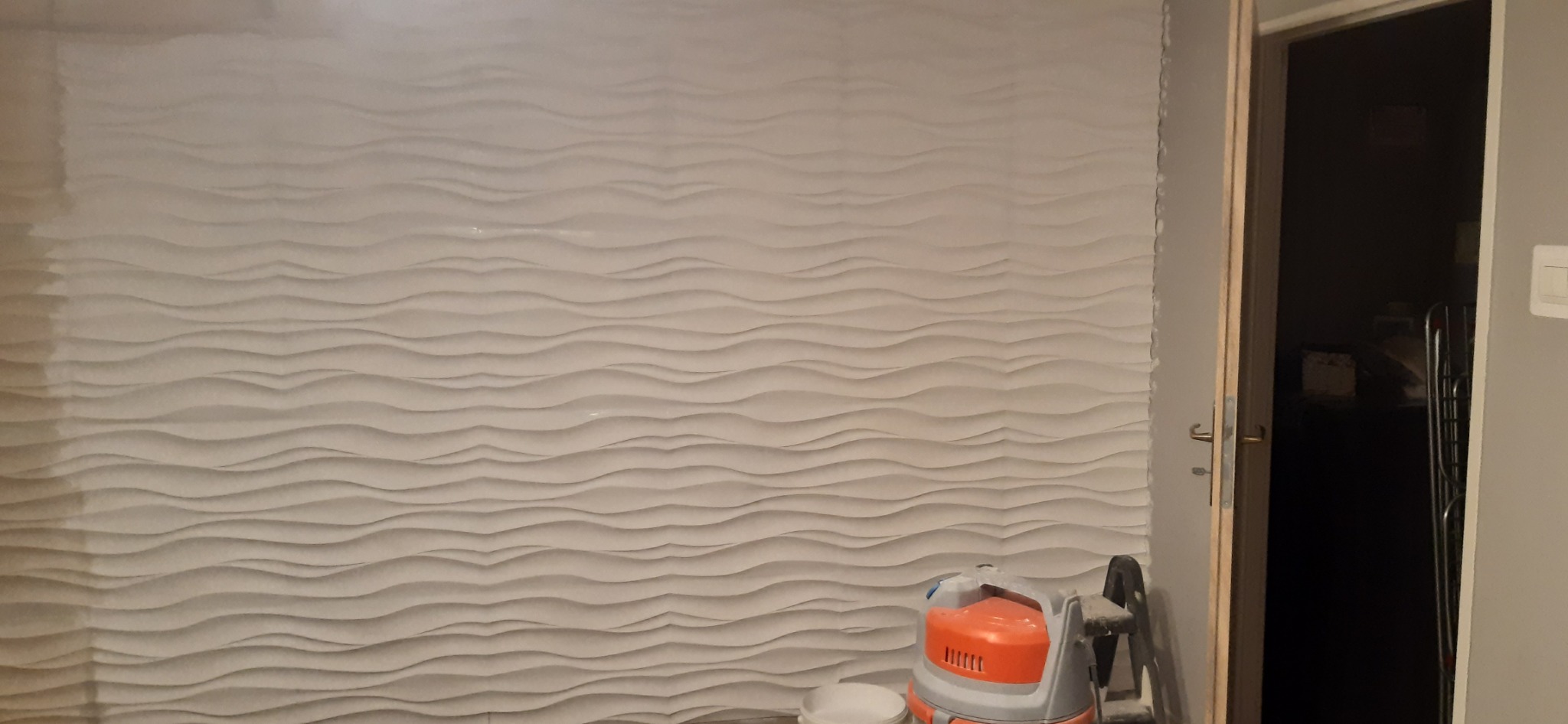 Ściana pokryta gładzią strukturalną w odcieniach bieli, z widocznym odkurzaczem budowlanym i otwartymi drzwiami do ciemnego pomieszczenia