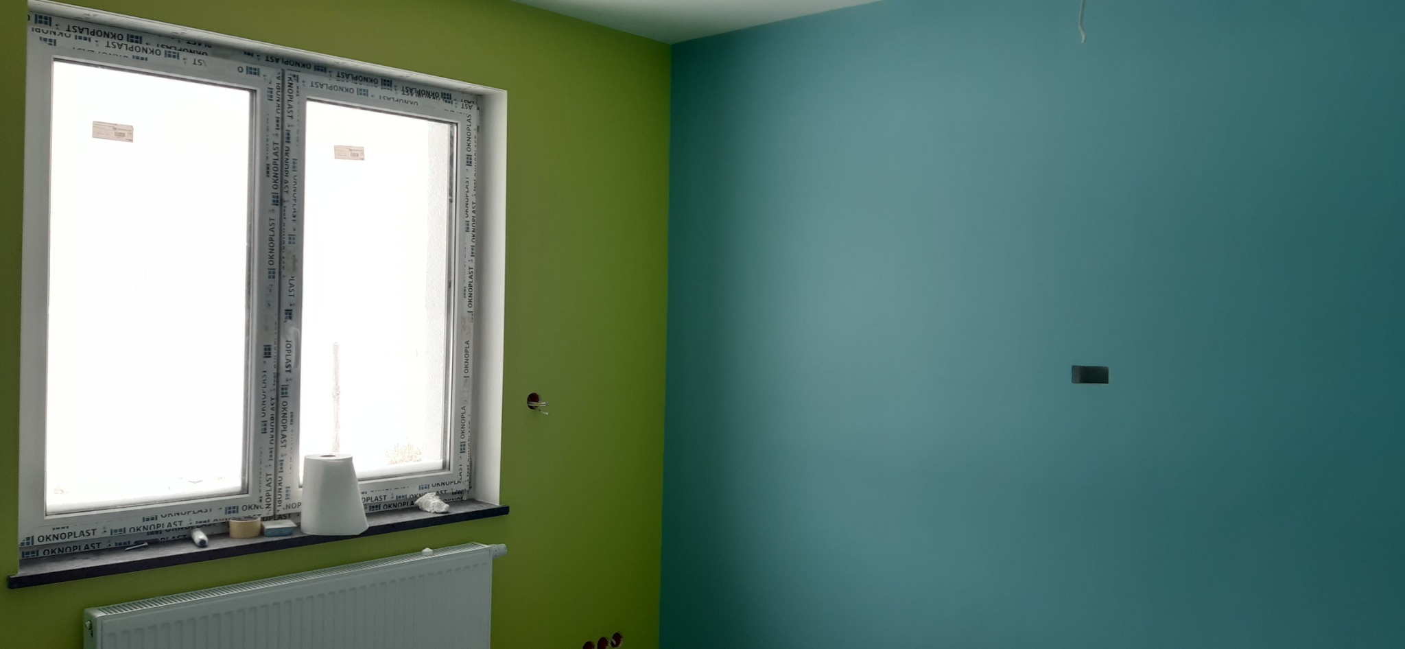 Pomalowane na zielono i turkusowo ściany pokoju z oknem PCV marki Oknoplast, na parapecie rolka papieru, grzejnik pod oknem, widoczne puszki elektryczne bez osprzętu.