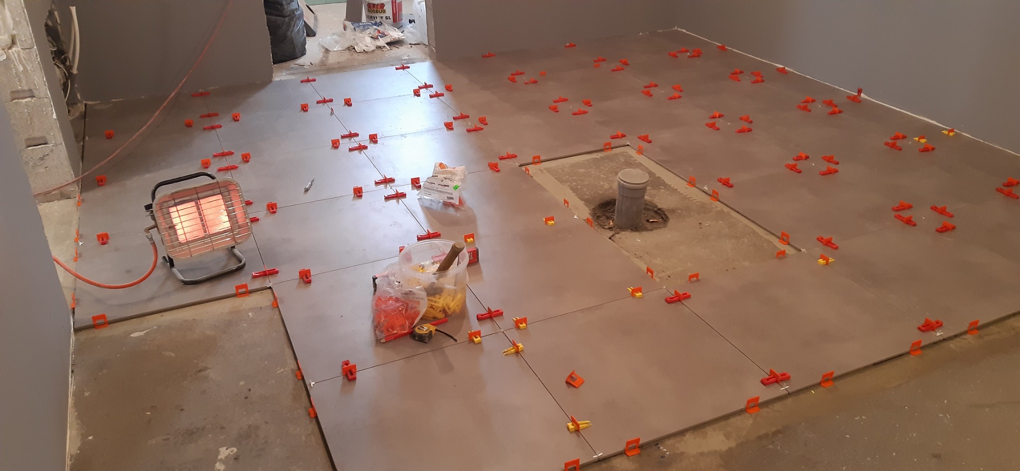 Układanie szarych płytek podłogowych z użyciem pomarańczowych klipsów poziomujących; widoczny grzejnik elektryczny, narzędzia i nieukończone fragmenty podłogi.