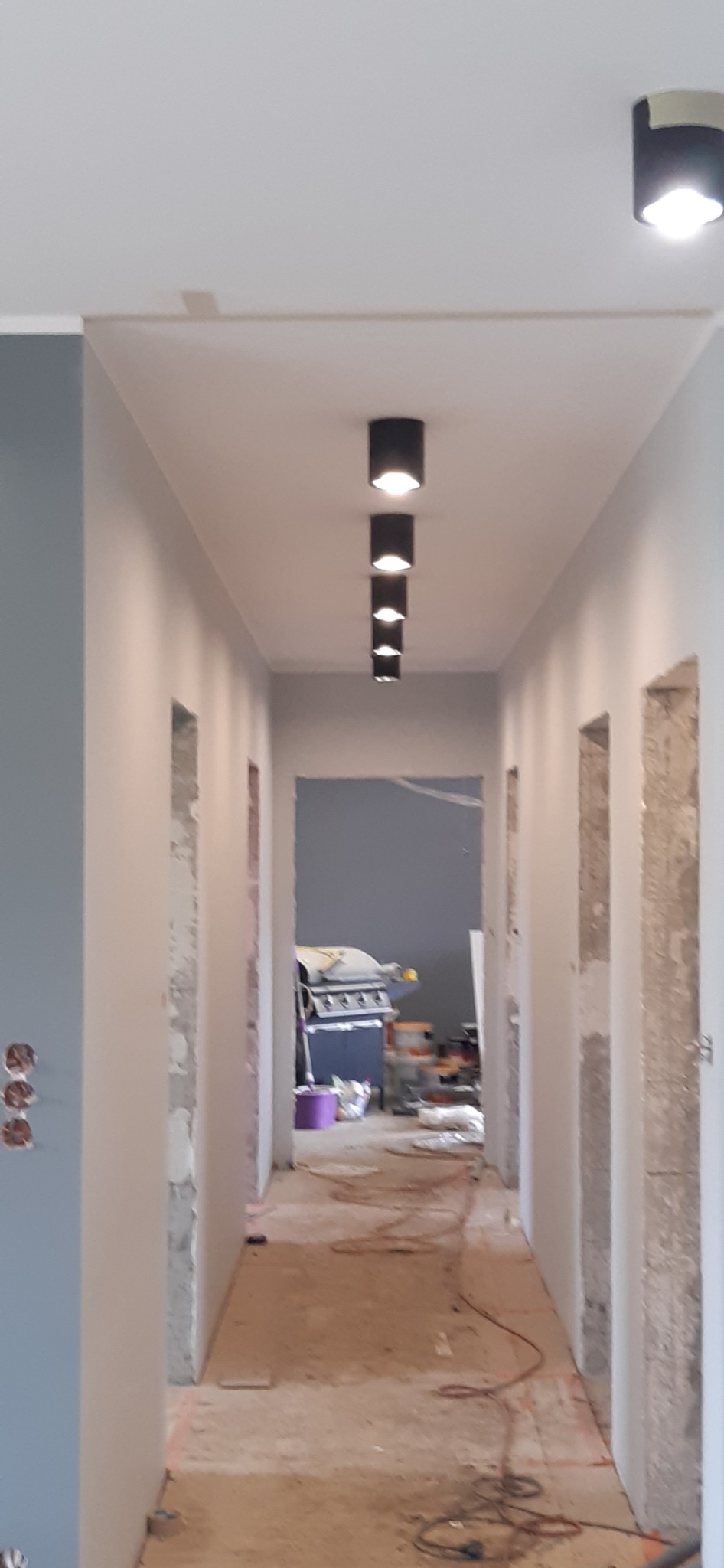 Korytarz w trakcie remontu z widocznymi surowymi ścianami, gładzią, wystającymi przewodami i rzędem czarnych lamp sufitowych