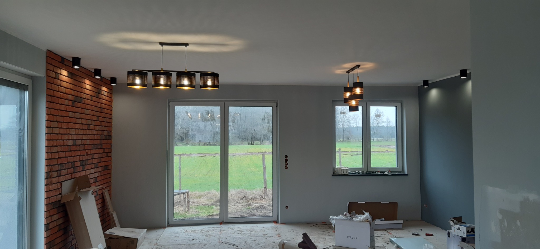 Wnętrze w trakcie wykańczania: ściana z cegły dekoracyjnej, szare ściany, dwa okna z widokiem na zewnątrz, nowoczesne lampy sufitowe i oświetlenie punktowe, podłoga zabezpieczona folią, widoczne...