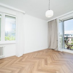 MASTER-UA NR 1 - Jasne wnętrze pokoju z parkietem w jodełkę, białymi ścianami, oknem z białymi firankami i balkonem z drewnianym stolikiem i krzesłami, widok na budynki za oknem.