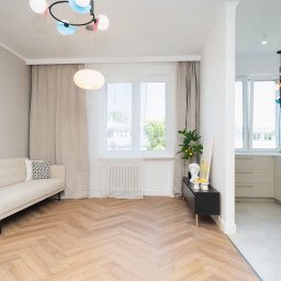 MASTER-UA NR 1 - Jasny salon z parkietem w jodełkę, kremową sofą, zasłonami i widokiem na kuchnię przez łuk w ścianie, urozmaicony kolorową lampą sufitową i dekoracjami.