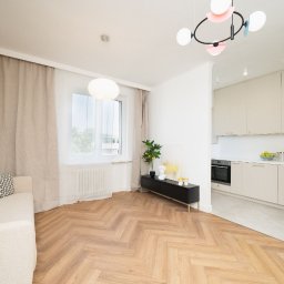 MASTER-UA NR 1 - Jasne, przestronne wnętrze po remoncie z parkietem ułożonym w jodełkę, beżową sofą z geometrycznymi poduszkami, czarną szafką RTV, widokiem na kuchnię i designerską lampą sufitową.