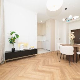 MASTER-UA NR 1 - Jasne wnętrze mieszkania z parkietem w jodełkę, okrągłym stołem z czterema krzesłami typu boucle, czarną szafką RTV z dekoracjami i widoczną kuchnią w tle.