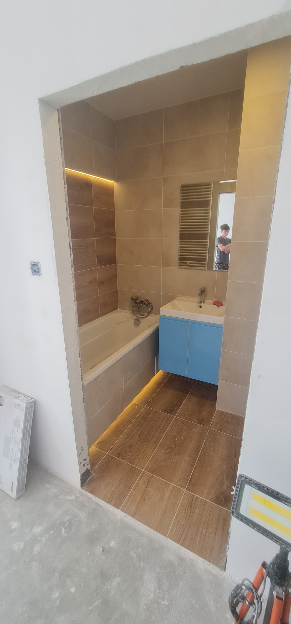 Wnętrze łazienki w trakcie remontu, widoczna wanna, umywalka z niebieską szafką, brązowe płytki podłogowe i ścienne, oświetlenie LED pod wanną i sufitem, surowy stan wykończenia przy wejściu.