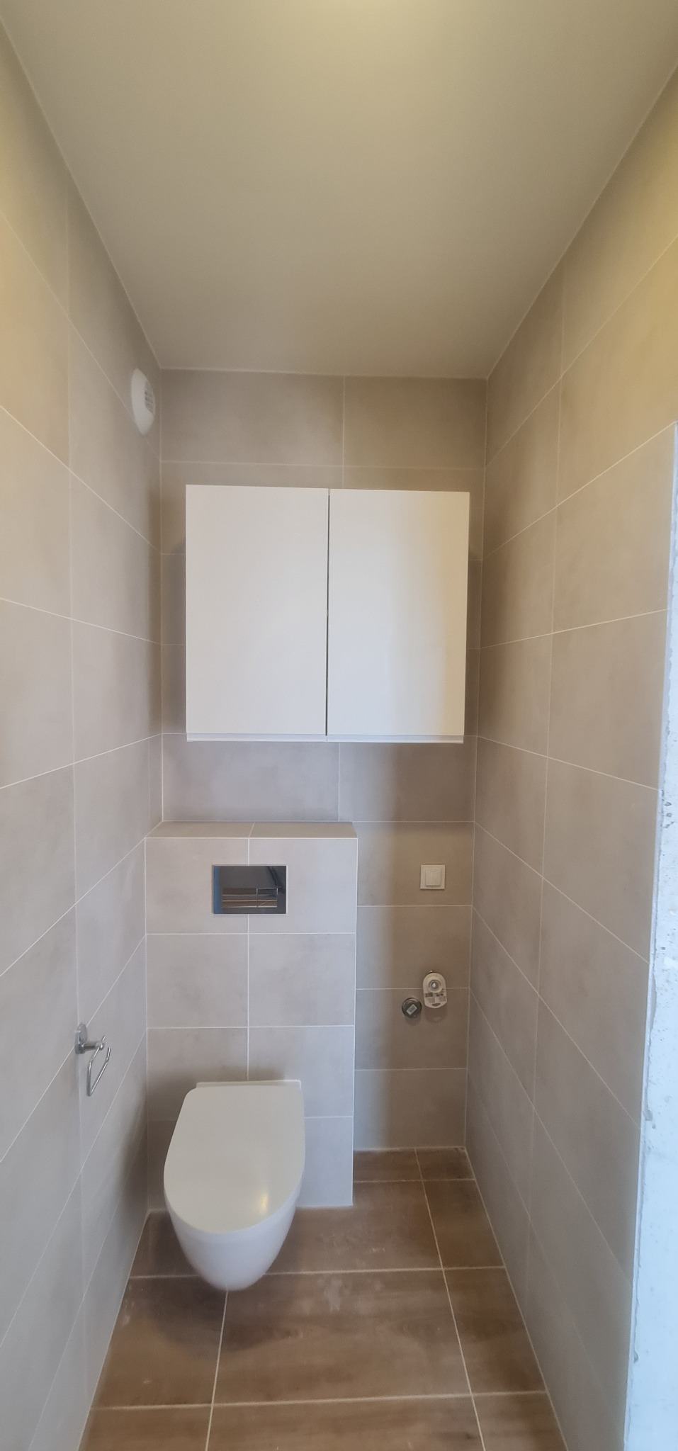 Zakończona aranżacja małej łazienki w Piasecznie z podwieszaną toaletą, szafką nad stelażem i beżowymi płytkami imitującymi kamień na ścianach i brązowymi na podłodze.