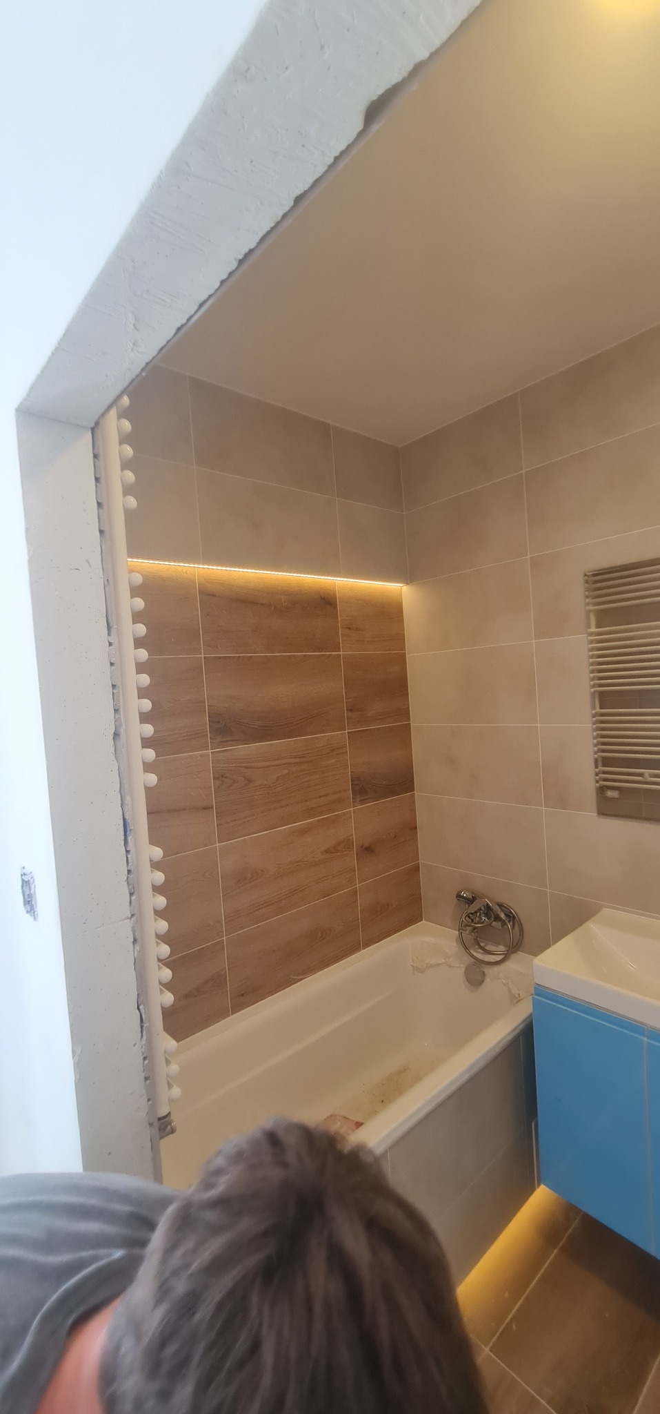 Remontowana łazienka z podświetlanymi płytkami ściennymi imitującymi drewno, białą wanną zabezpieczoną folią i niebieską szafką umywalkową z podświetleniem LED u dołu. Na ścianie widoczny grzejnik...