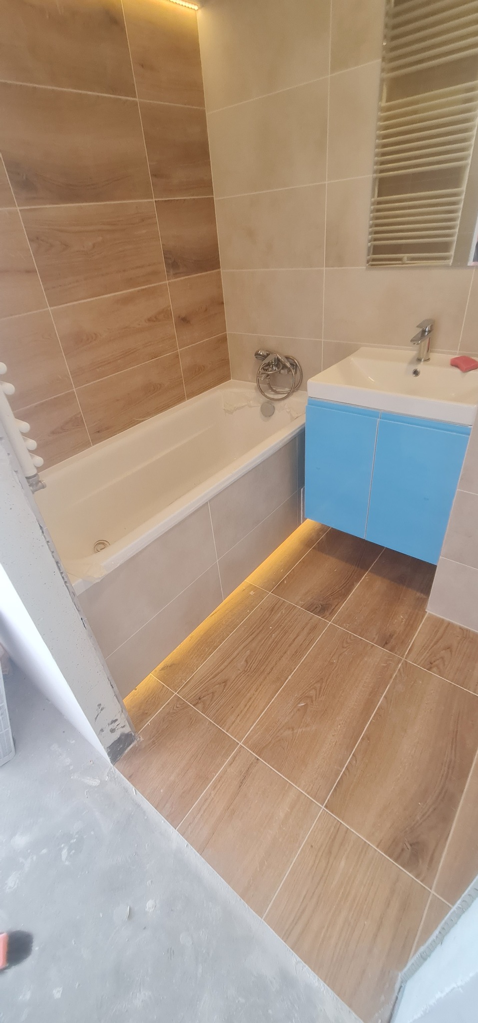 Wykończona łazienka z beżowymi i brązowymi płytkami imitującymi drewno, podświetleniem LED pod wanną i szafką umywalkową w kolorze błękitnym.