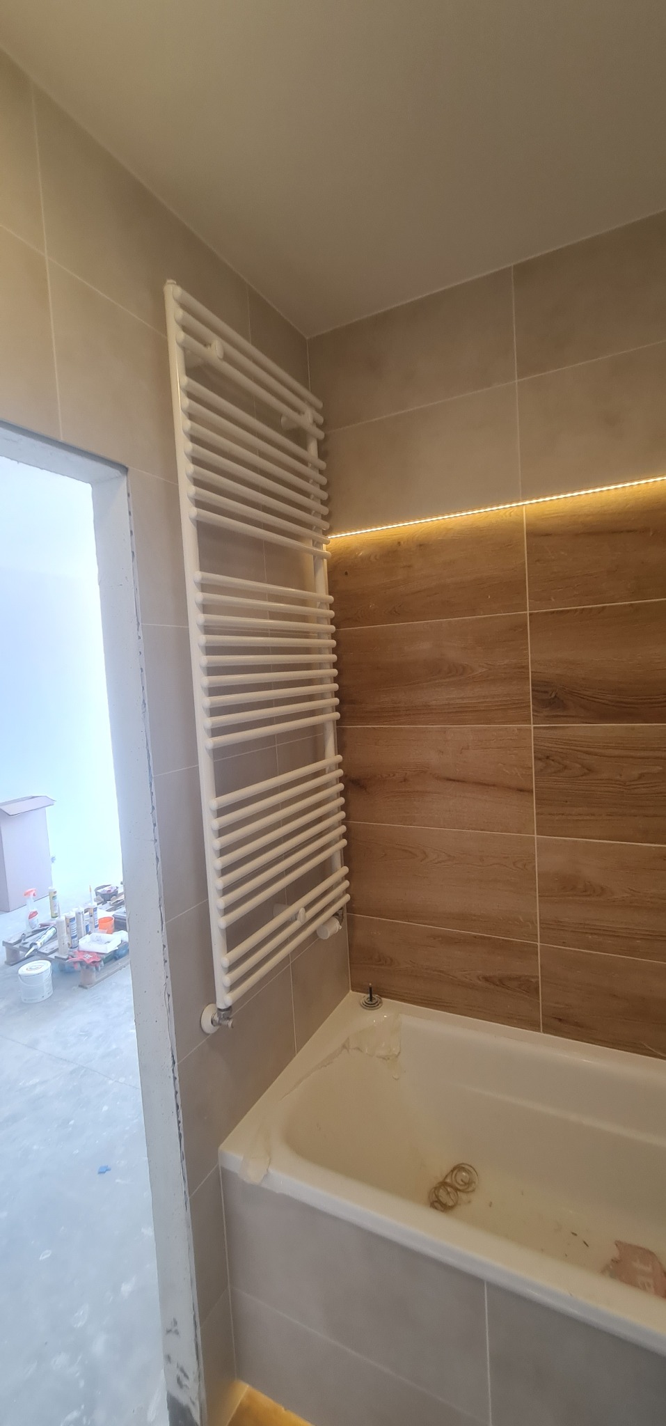 Biały grzejnik drabinkowy zamontowany na ścianie w łazience z brązowymi płytkami imitującymi drewno i podświetleniem LED, widok na wannę z folią ochronną i remontowane pomieszczenie przez otwarte...