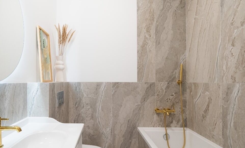 Nowoczesna łazienka z marmurowymi płytkami, złotą armaturą, okrągłym lustrem i dekoracyjnym wazonem z trawami na półce.