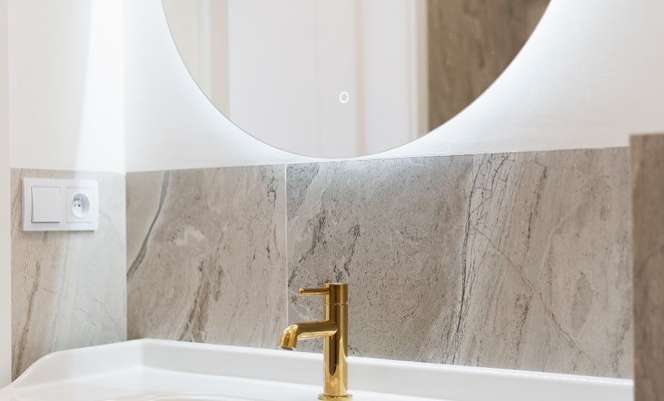 Nowoczesna łazienka z okrągłym lustrem podświetlanym LED, złotą baterią umywalkową i szarymi płytkami imitującymi marmur.