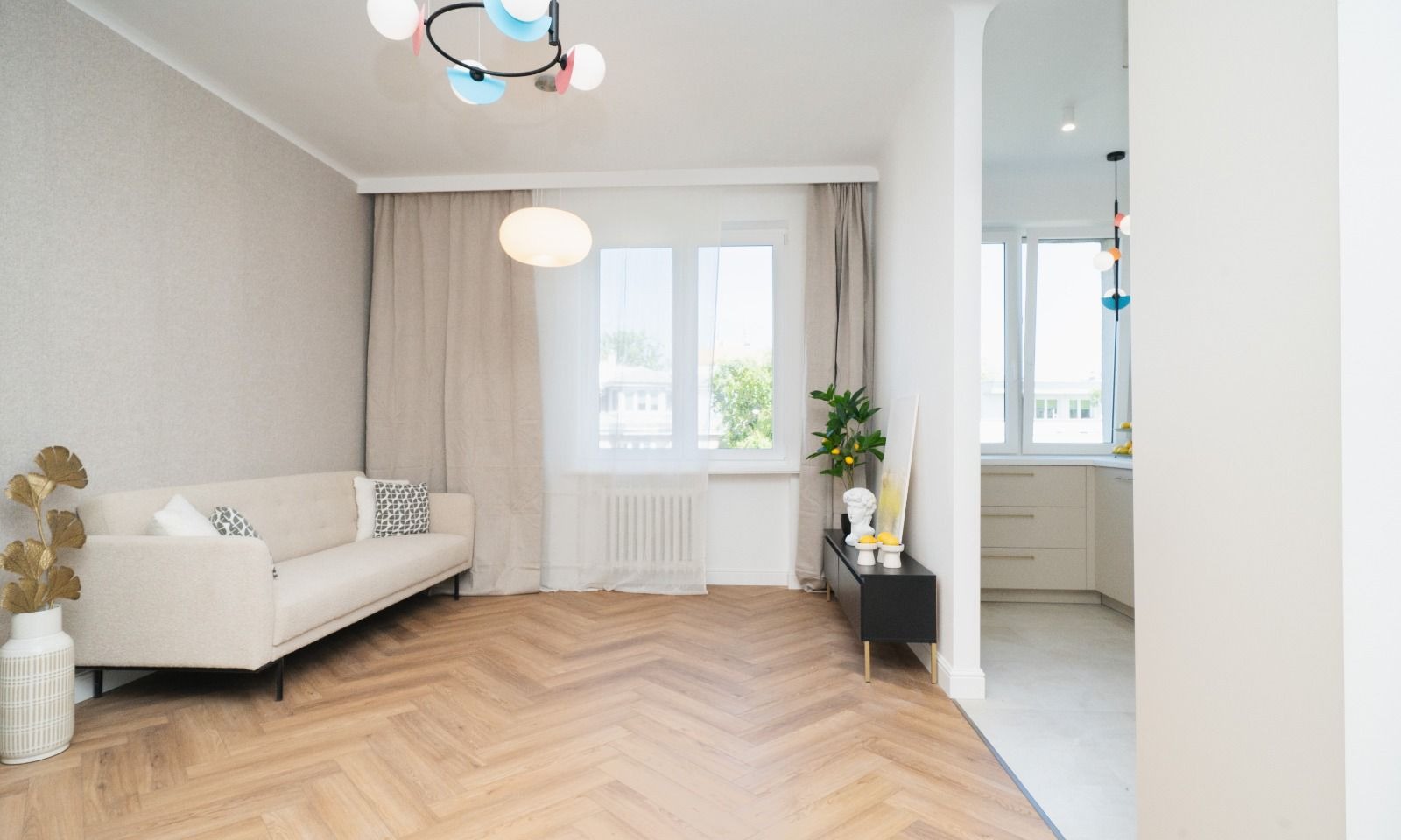 Jasny salon z parkietem w jodełkę, kremową sofą, zasłonami i widokiem na kuchnię przez łuk w ścianie, urozmaicony kolorową lampą sufitową i dekoracjami.