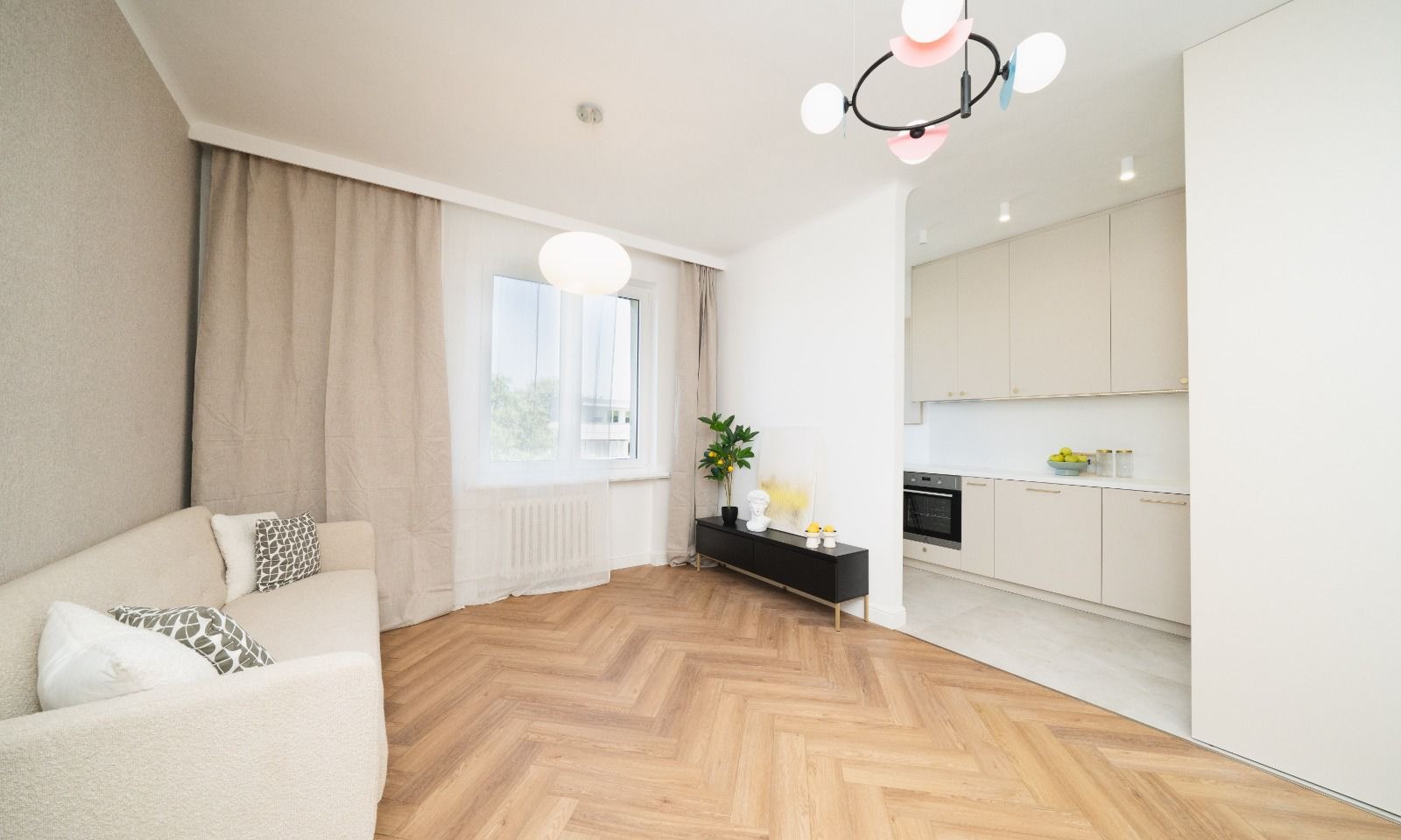 Jasne, przestronne wnętrze po remoncie z parkietem ułożonym w jodełkę, beżową sofą z geometrycznymi poduszkami, czarną szafką RTV, widokiem na kuchnię i designerską lampą sufitową.