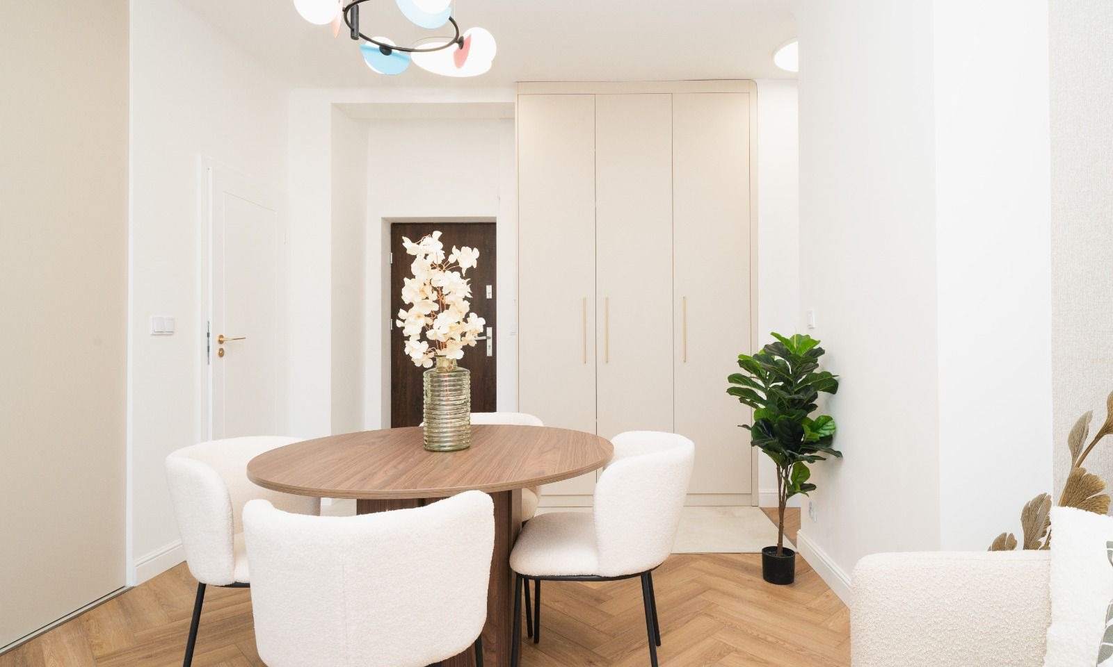 Jasne wnętrze jadalni z okrągłym stołem z drewna, białymi krzesłami typu bouclé, dużą szafą wnękową i parkietem ułożonym w jodełkę.