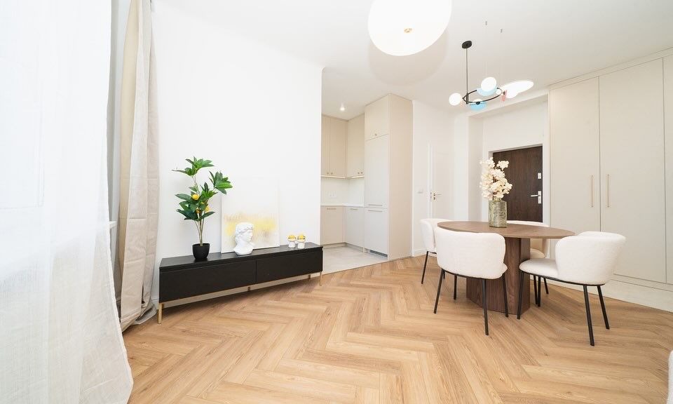 Jasne wnętrze mieszkania z parkietem w jodełkę, okrągłym stołem z krzesłami, czarną komodą z dekoracjami i widokiem na kuchnię w tle.