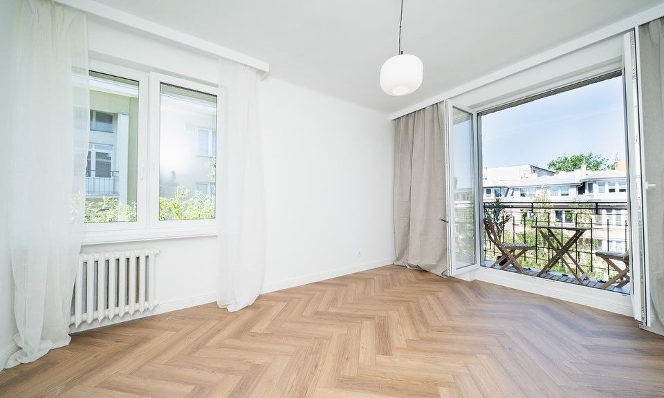 Jasne, puste wnętrze z parkietem w jodełkę, białymi ścianami, grzejnikiem pod oknem i balkonem z drewnianym stolikiem i krzesłami widocznym przez otwarte drzwi balkonowe.