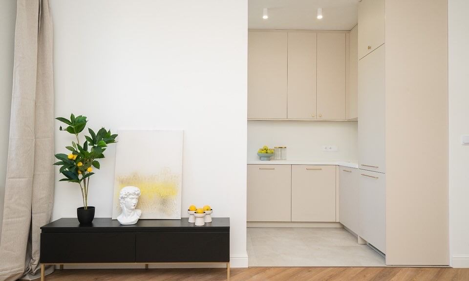 Jasne wnętrze mieszkania z elementami dekoracyjnymi: czarna komoda z rośliną cytrynową, popiersie i obraz na tle beżowej zabudowy kuchennej.