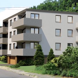 KRYJAK Biuro Projektów - Wizualizacja nowoczesnego, trzypiętrowego budynku mieszkalnego z balkonami, otoczonego zielenią i z zaparkowanym samochodem na ulicy.