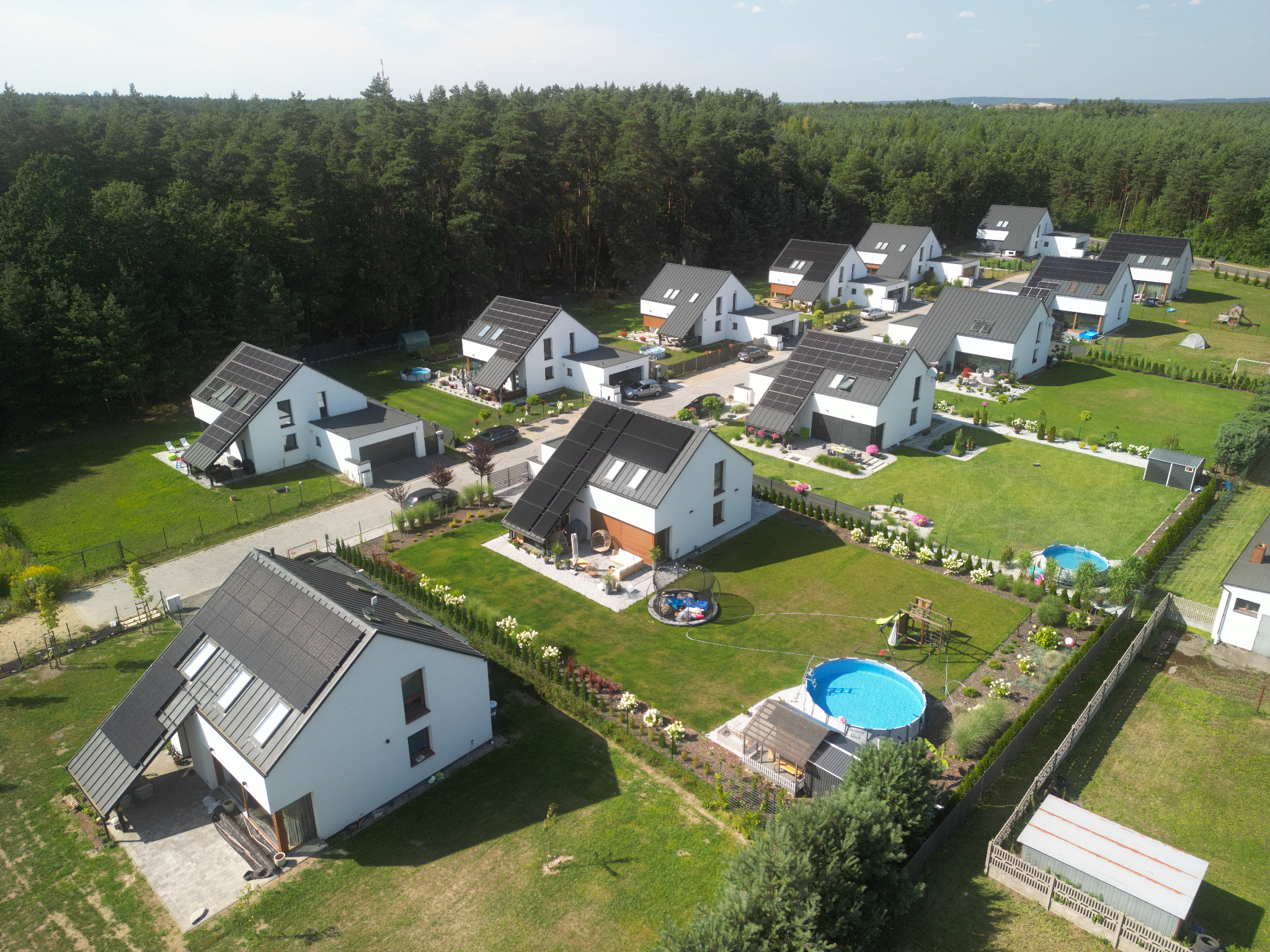 Osiedle nowoczesnych, parterowych domów jednorodzinnych z panelami fotowoltaicznymi na dachach, widok z lotu ptaka, z zadbanymi ogrodami i basenami na trawnikach, otoczone lasem.