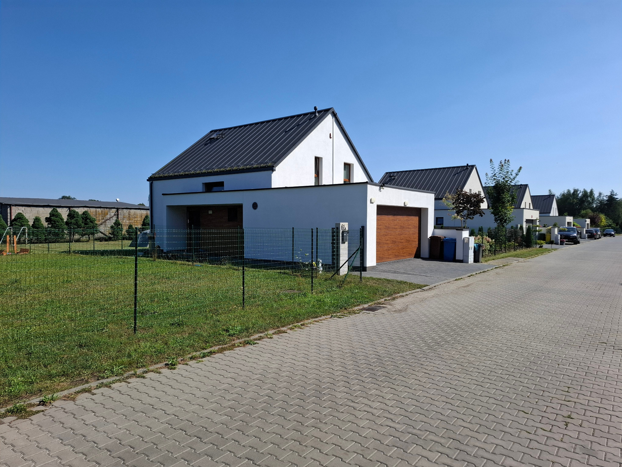 Nowoczesne, białe domy jednorodzinne z ciemnymi dachami, widok z ulicy w słoneczny dzień, z zielonym trawnikiem i ogrodzeniem.