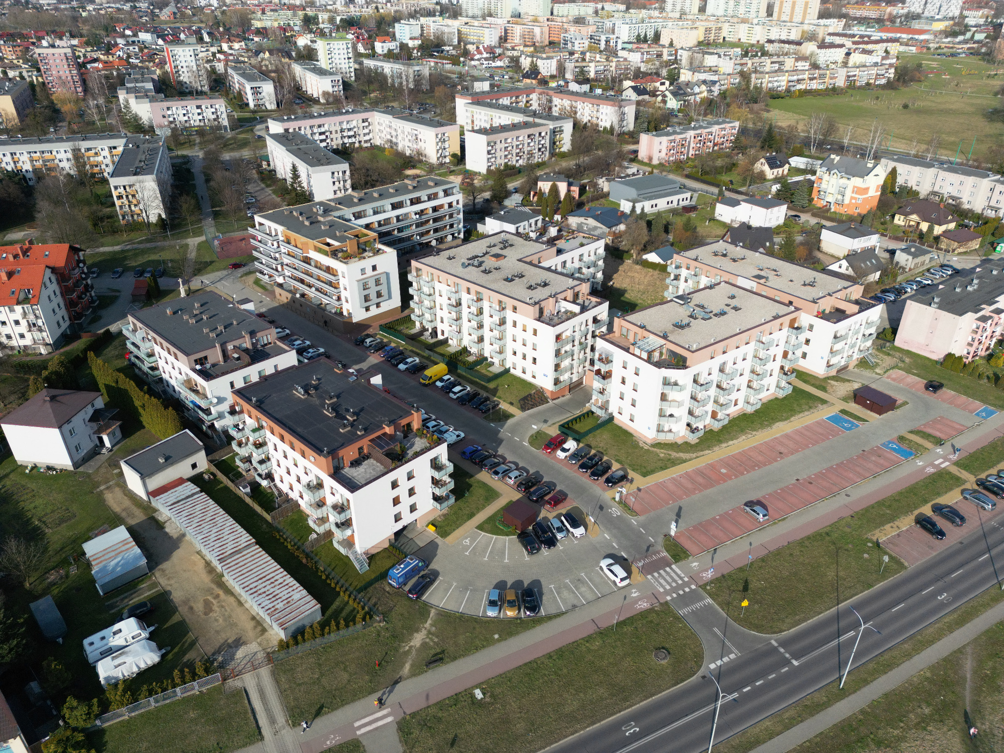Widok z lotu ptaka na kompleks nowoczesnych budynków mieszkalnych z balkonami, z parkingiem i drogą dojazdową, otoczonych zielenią i zabudową miejską w tle.