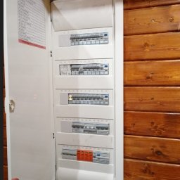 Leszek Lendzion elektryk - Otwarta skrzynka rozdzielcza z wyłącznikami na tle drewnianej ściany, widoczna lista obwodów na wewnętrznej stronie drzwiczek.