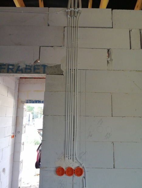 Surowe wnętrze w trakcie budowy: białe bloczki, widoczne kable elektryczne w białych rurkach prowadzone pionowo wzdłuż ściany, pomarańczowe puszki podtynkowe. Częściowo widoczny otwór drzwiowy...