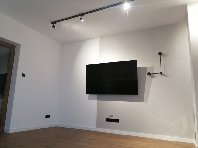Minimalistyczny salon z zamontowanym telewizorem, czarną listwą oświetleniową sufitową z reflektorami i dekoracją ścienną w kształcie geometrycznym.