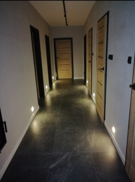 Oświetlony korytarz z ciemną podłogą i punktowymi lampami w ścianach, widoczne drzwi z jasnego drewna z czarnymi ościeżnicami i czarna szyna oświetleniowa na suficie.