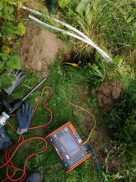 Pomiar rezystancji uziomu w Toruniu: Miernik, przewody pomiarowe, elektroda wbita w ziemię, obok leżą rękawice robocze i młot udarowy, widoczne kable w peszlu.