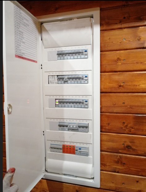 Otwarta skrzynka rozdzielcza z wyłącznikami na tle drewnianej ściany, widoczna lista obwodów na wewnętrznej stronie drzwiczek.