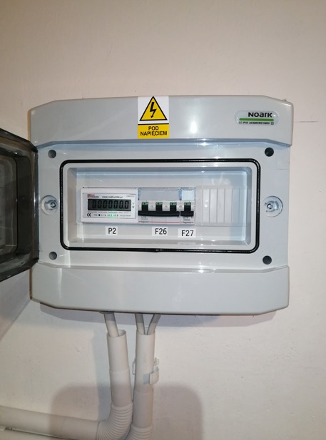 Rozdzielnica elektryczna Noark z otwartymi drzwiczkami, widoczne zabezpieczenia F26 i F27 oraz licznik energii z wyświetlaczem LCD, żółta naklejka ostrzegawcza 'Pod Napięciem', przewody w peszlach.