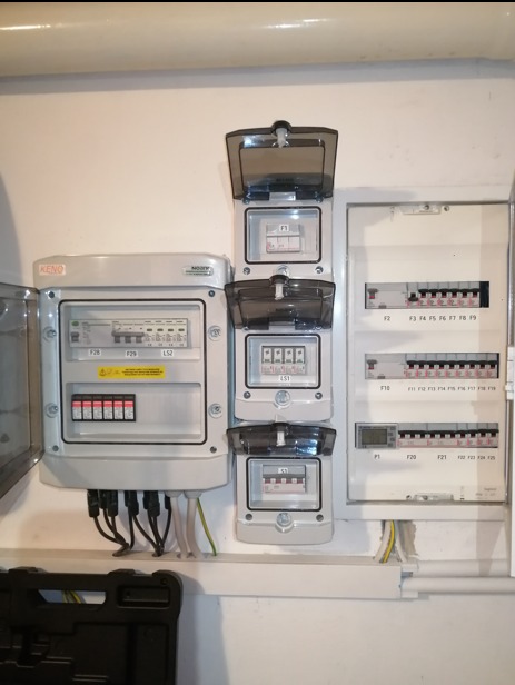 Rozdzielnica elektryczna z trzema licznikami w obudowach z uchylnymi klapkami, obok skrzynka z bezpiecznikami i przewodami. Widoczna instalacja elektryczna na białej ścianie.