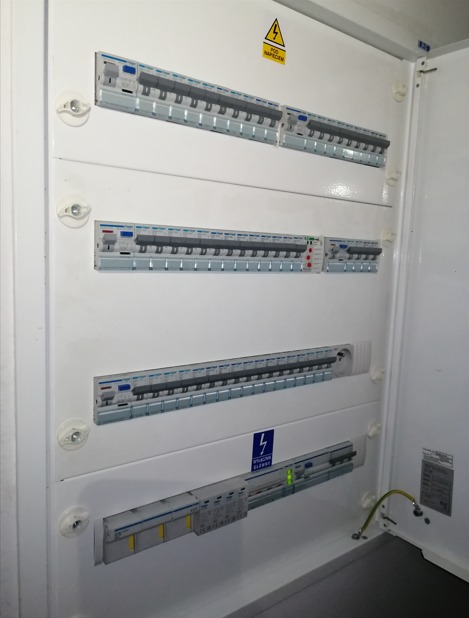 Otwarta, biała skrzynka rozdzielcza z zabezpieczeniami różnicowoprądowymi i nadprądowymi, widoczne przewody ochronne, naklejka ostrzegawcza o napięciu.