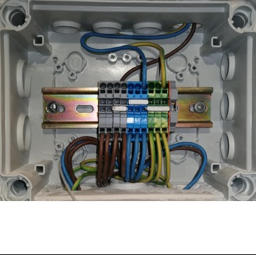 Wnętrze puszki elektrycznej z zamontowanymi złączkami listwowymi na szynie DIN, widoczne przewody w różnych kolorach (niebieski, zielono-żółty, brązowy), całość w szarej obudowie z widocznymi...