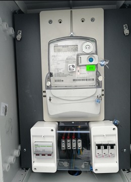 Zamknięta szafka elektryczna z zamontowanym licznikiem energii elektrycznej i zabezpieczeniami nadprądowymi, widoczne przewody w izolacji, szara obudowa szafki.