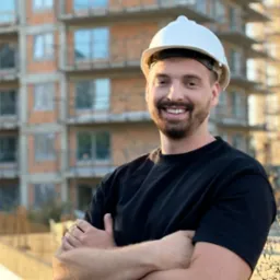 Uśmiechnięty mężczyzna w kasku ochronnym na tle budynku w stanie surowym, skrzyżowane ręce, ciepłe światło popołudniowe.