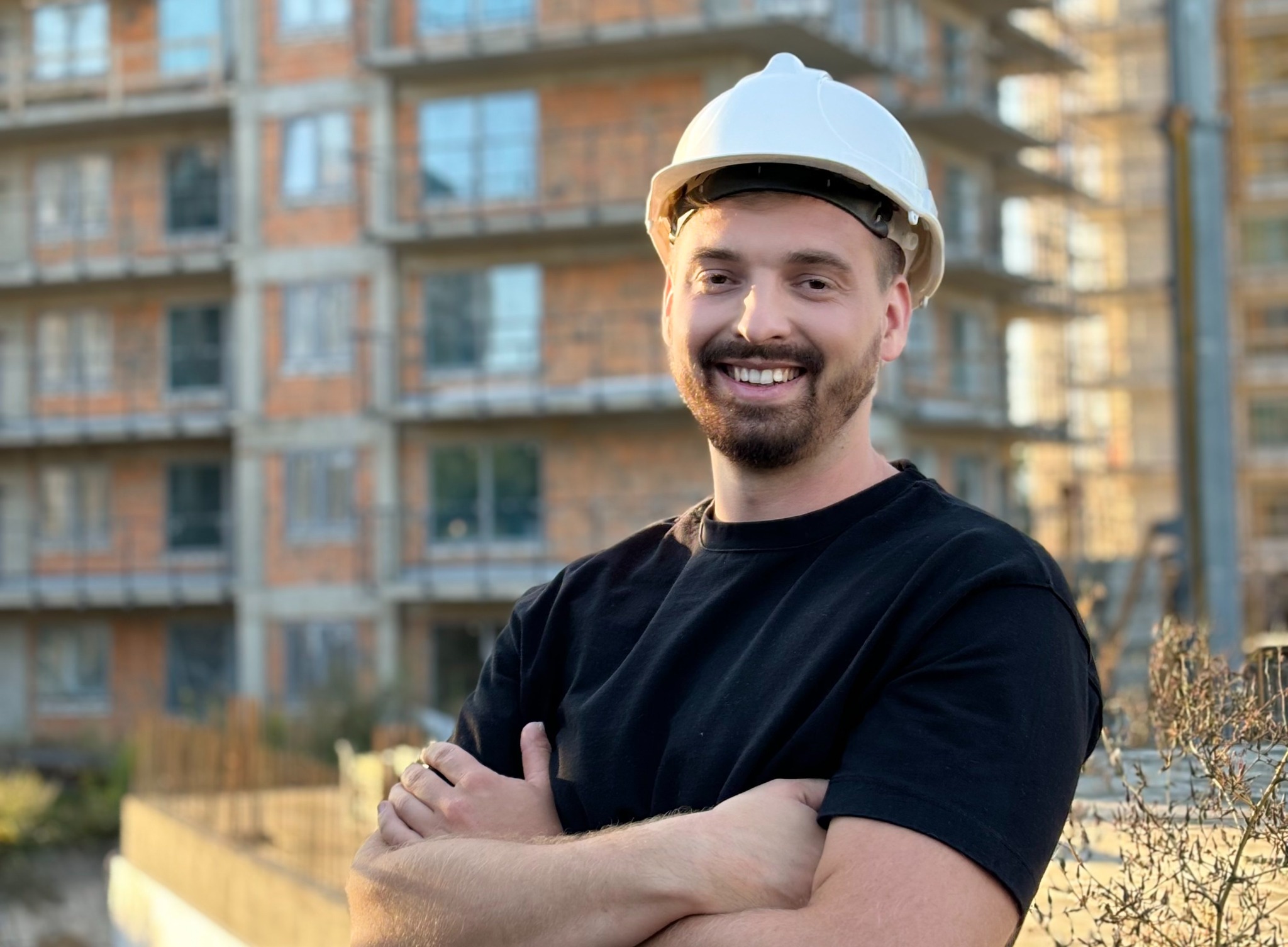 Uśmiechnięty mężczyzna w kasku ochronnym na tle budynku w stanie surowym, skrzyżowane ręce, ciepłe światło popołudniowe.