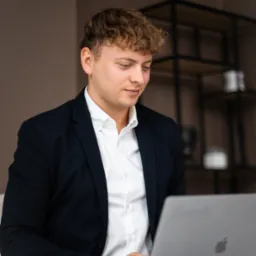 Młody mężczyzna w garniturze i białej koszuli, z kręconymi włosami, siedzi i pracuje na laptopie, w tle regał w stylu industrialnym.