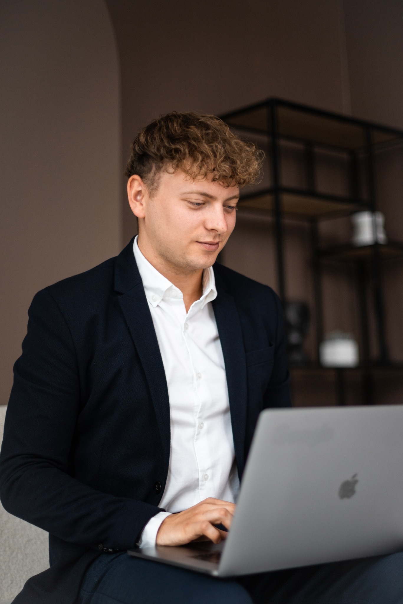 Młody mężczyzna w garniturze i białej koszuli, z kręconymi włosami, siedzi i pracuje na laptopie, w tle regał w stylu industrialnym.